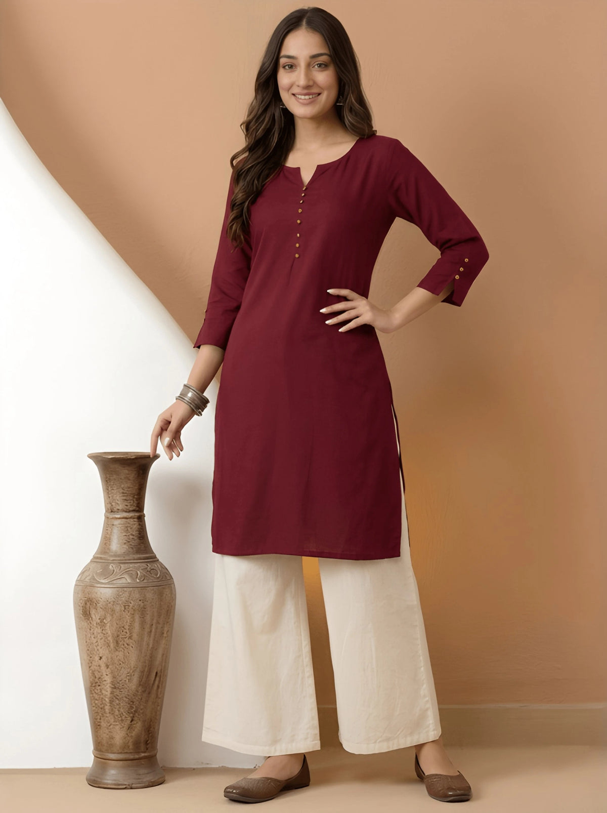 Woman wearing a maroon kurta with white pants standing next to a decorative vase against a brown background