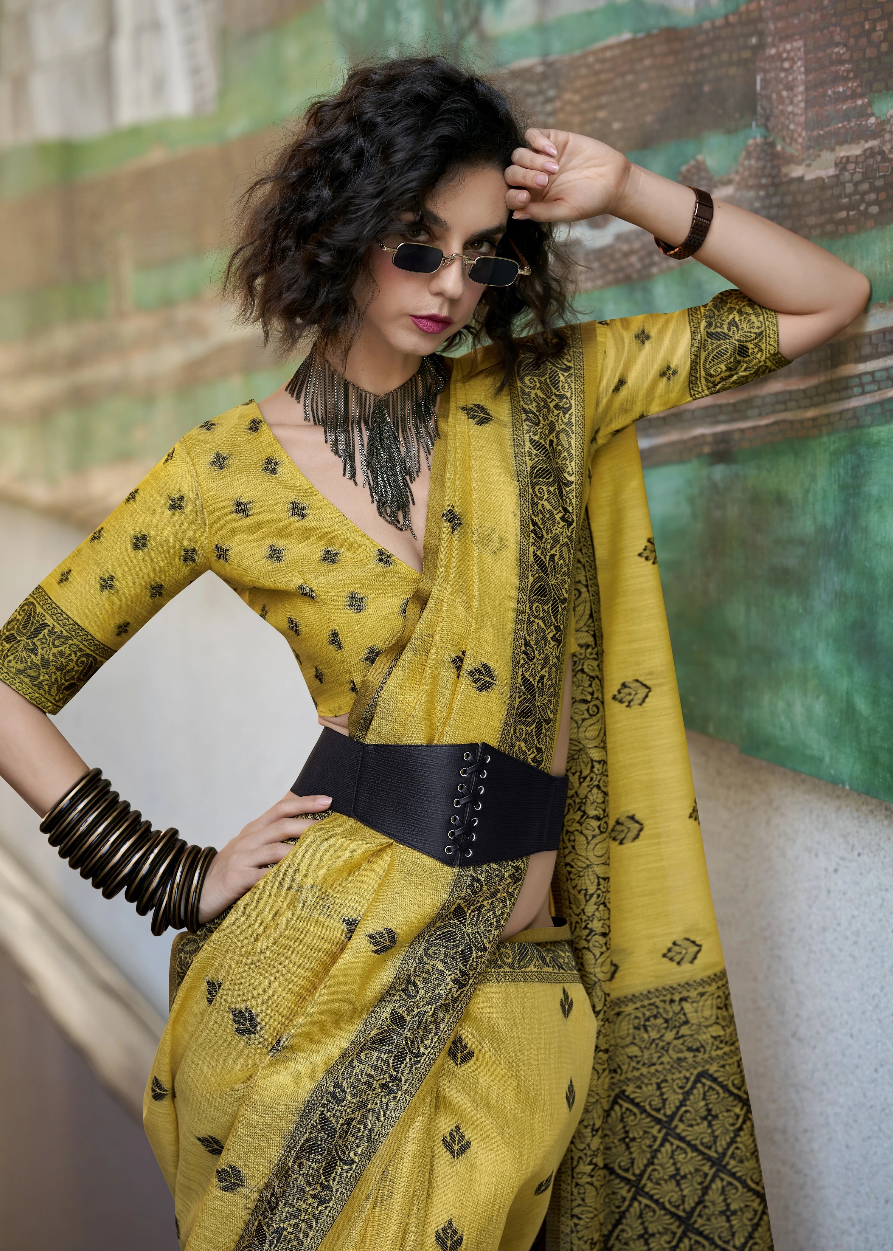 Woman wearing a yellow saree with black patterns against a textured wall.