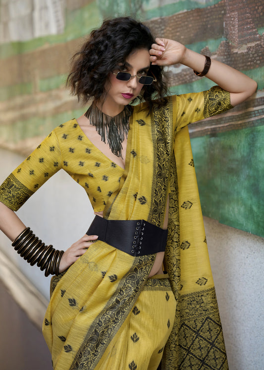 Woman wearing a yellow saree with black patterns against a textured wall.