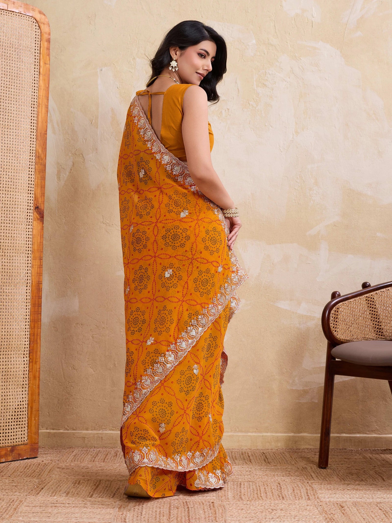 Woman wearing a bright orange saree with a beige wall and chair in the background