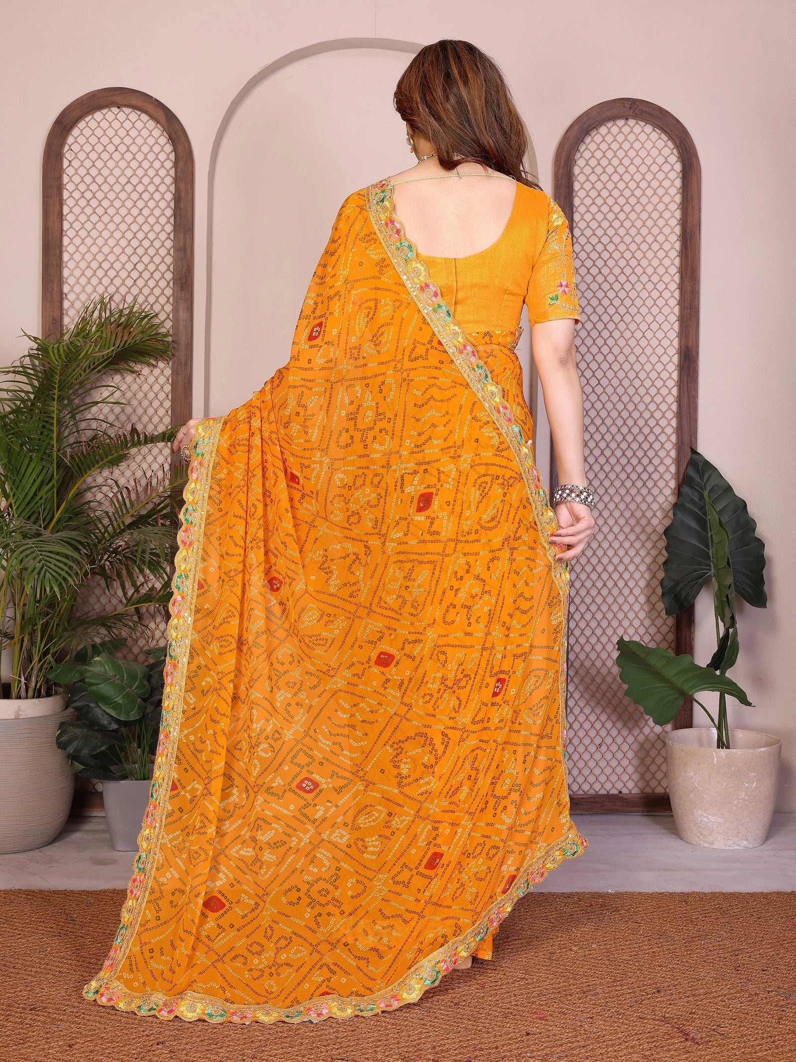 Woman wearing a yellow saree with a pattern, standing in a room with plants.
