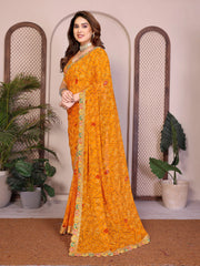 Woman wearing a bright orange saree with a floral pattern in a decorative setting with plants.