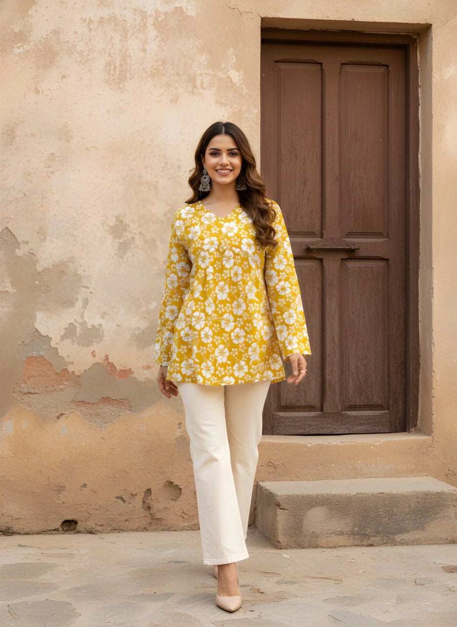 A full-length front view of the woman standing in a courtyard. She is wearing the mustard floral kurti paired with off-white wide-leg trousers and beige heels, set against a rustic beige wall and a dark brown wooden door.