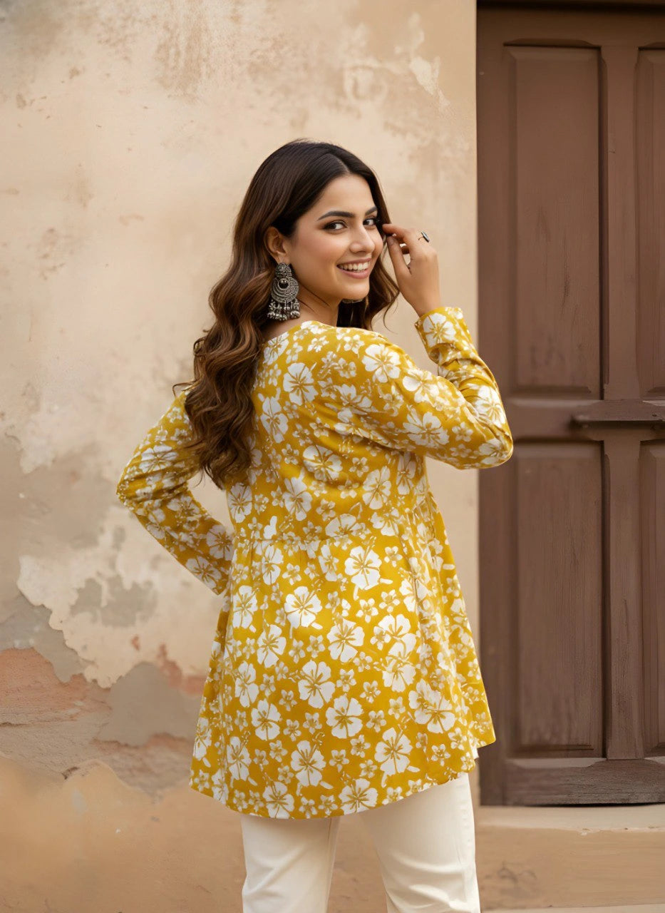 A woman is captured from behind, looking back over her shoulder with a bright smile. She is wearing a mustard-yellow short kurti with an all-over white hibiscus print and a gathered waistline that creates a soft flare.
