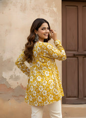A woman is captured from behind, looking back over her shoulder with a bright smile. She is wearing a mustard-yellow short kurti with an all-over white hibiscus print and a gathered waistline that creates a soft flare.
