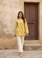 A full-length front view of the woman standing in a courtyard. She is wearing the mustard floral kurti paired with off-white wide-leg trousers and beige heels, set against a rustic beige wall and a dark brown wooden door.