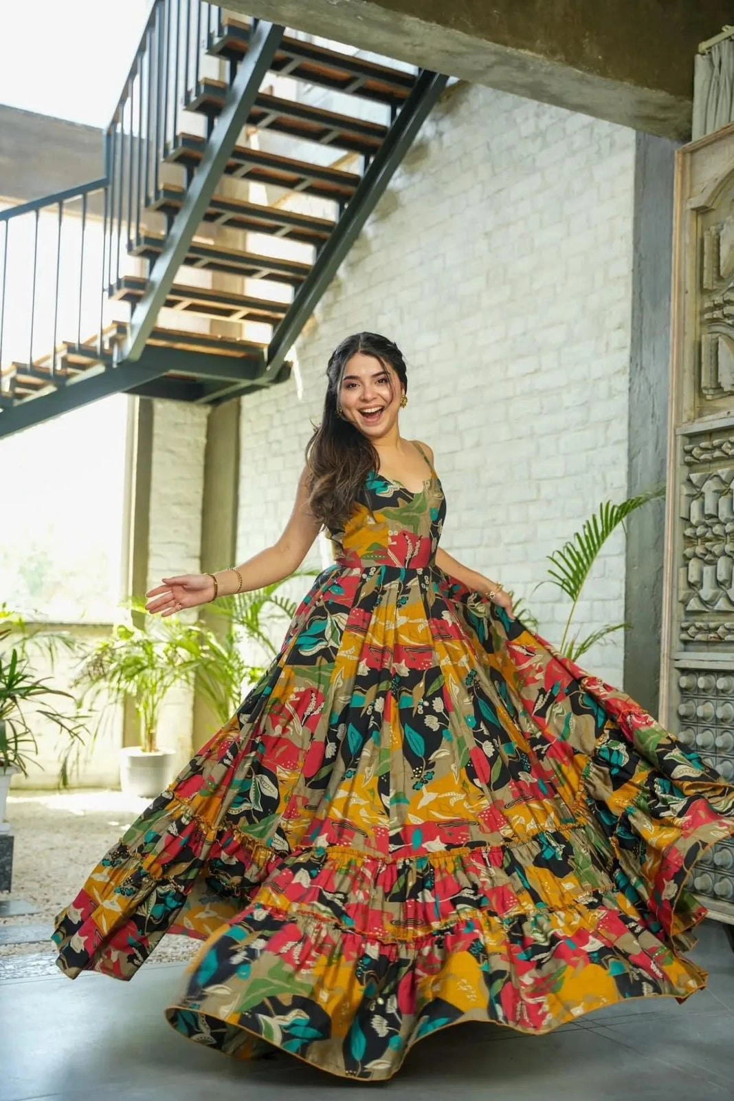 Woman in a colorful floral dress posing in an indoor setting with plants and stairs.