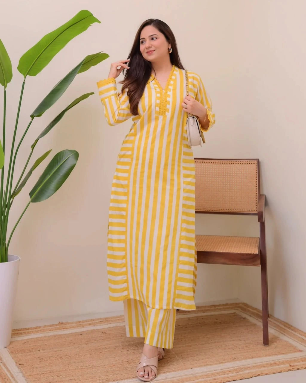 Woman in a yellow and white striped outfit standing indoors with a plant and chair in the background.