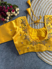 A bright yellow raw silk blouse with a deep U-neckline at the back. The edges are decorated with intricate gold and pearl floral embroidery featuring small green leaf accents. It includes matching yellow tie-back cords with double-layered triangular tassels, and small pearl buttis are scattered across the fabric.
