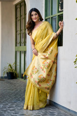 Woman in a yellow saree with floral patterns standing against a white wall.