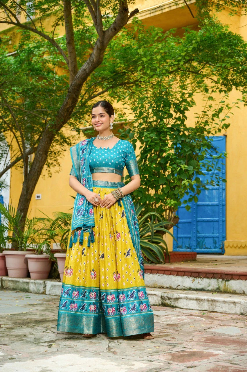 Woman in a yellow and blue traditional outfit standing outdoors with greenery and a yellow building in the background.