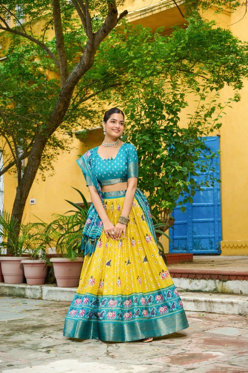 Woman in a traditional yellow and blue outfit standing outdoors with greenery and a yellow wall in the background.