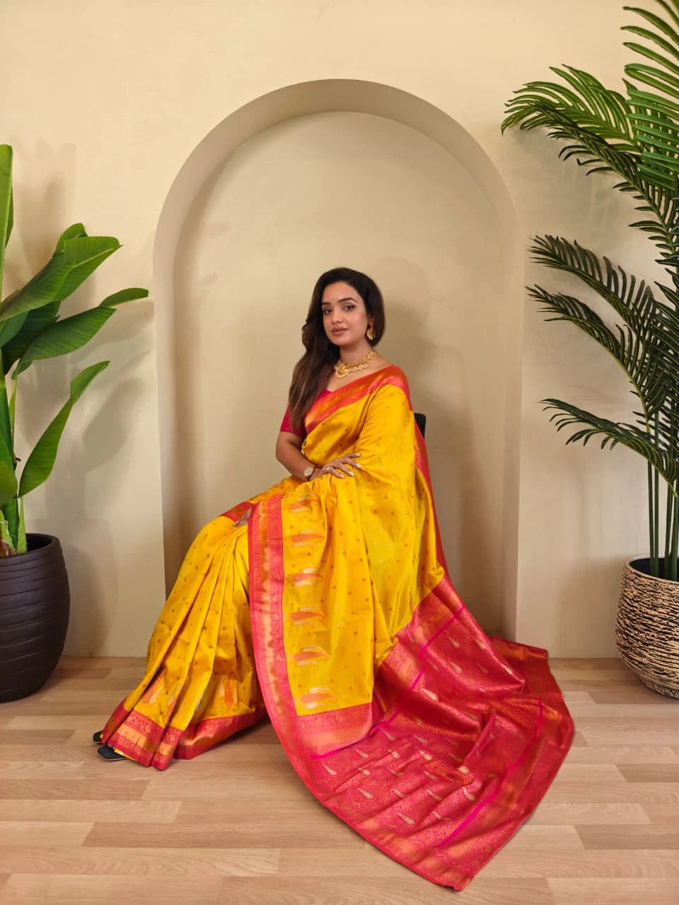 Woman in a yellow and pink saree sitting in a room with plants.