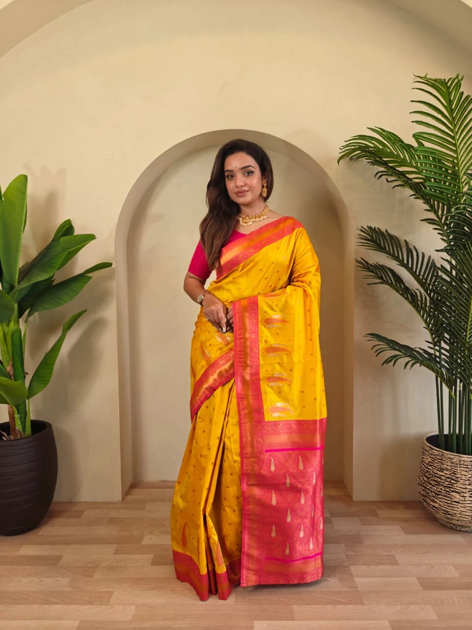 Woman in a yellow and pink saree standing in a room with plants.
