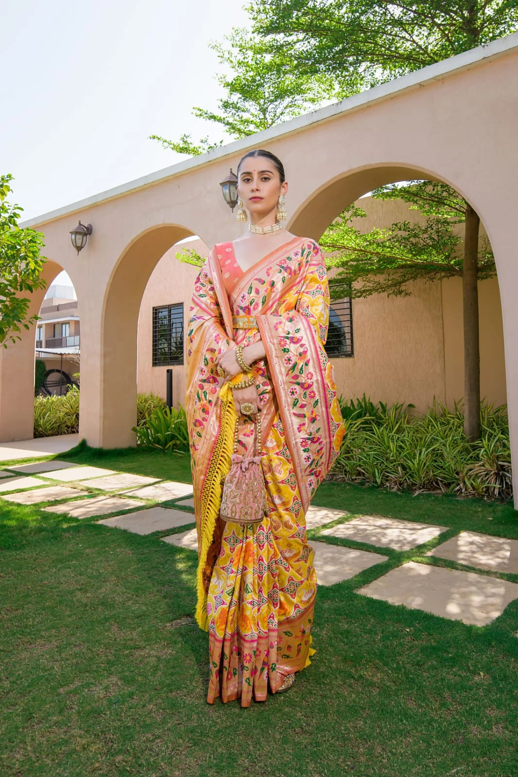 Woman in a colorful saree standing outdoors with greenery and a building in the background