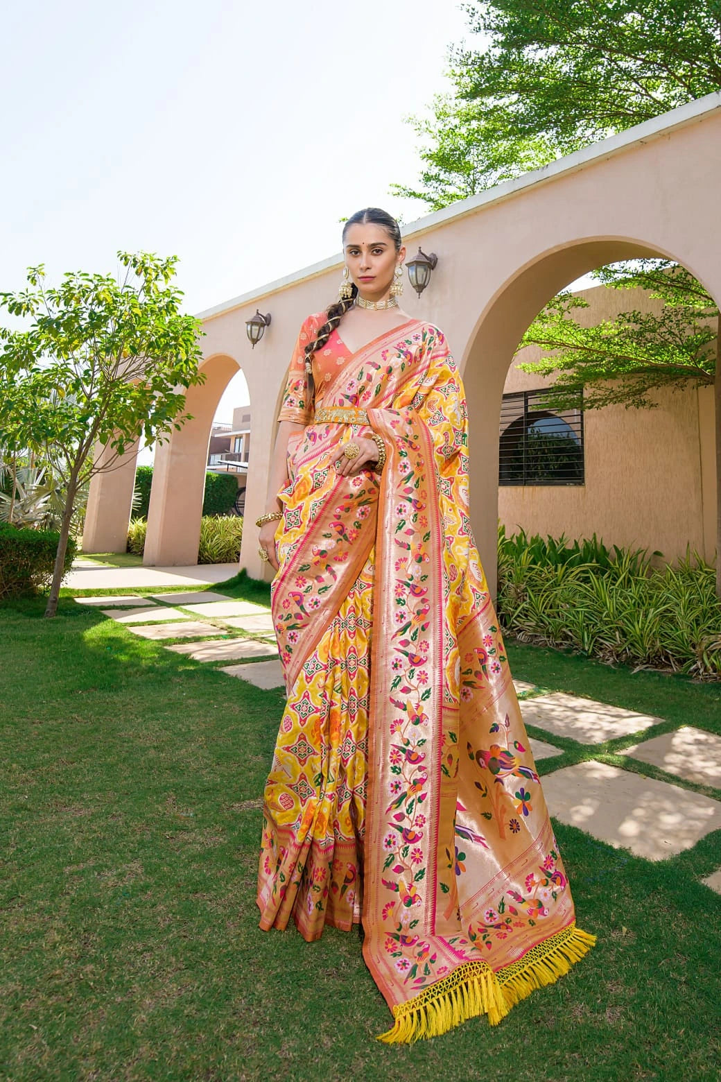Woman wearing a colorful saree outdoors with greenery and a building in the background