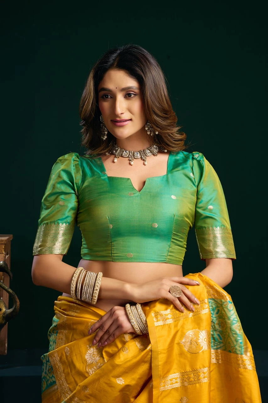 Woman wearing a green blouse and yellow saree with gold jewelry against a dark background