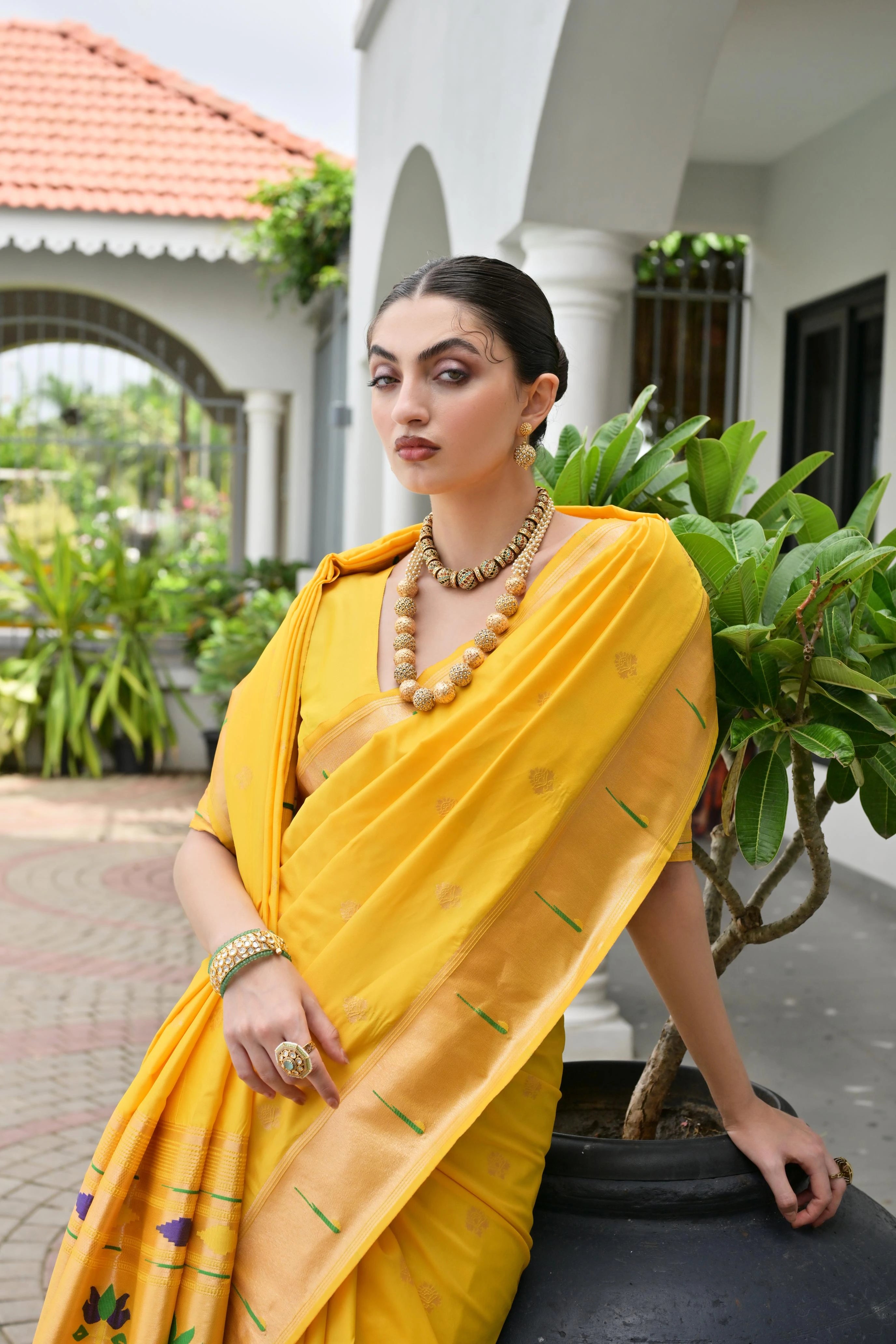 Woman in a yellow saree sitting outdoors with greenery and a building in the background