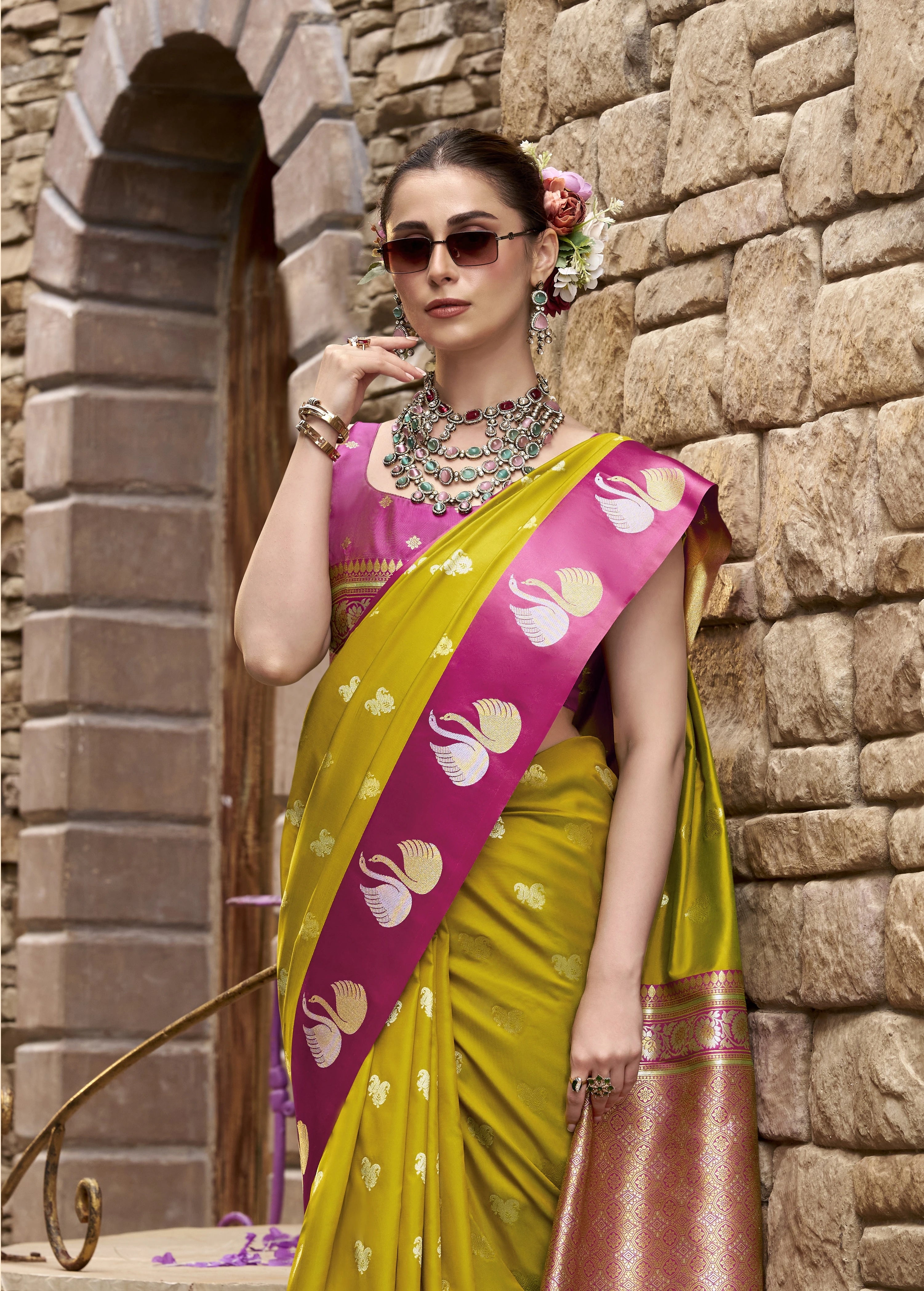 Woman in a yellow and pink saree standing against a stone wall.