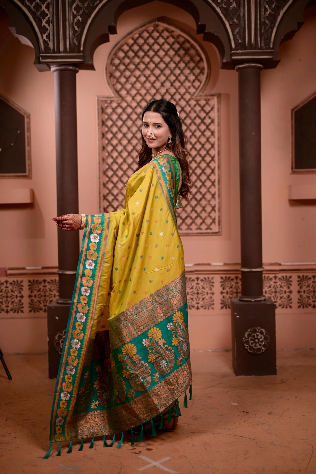 Woman in a yellow and green saree standing in an ornate indoor setting.