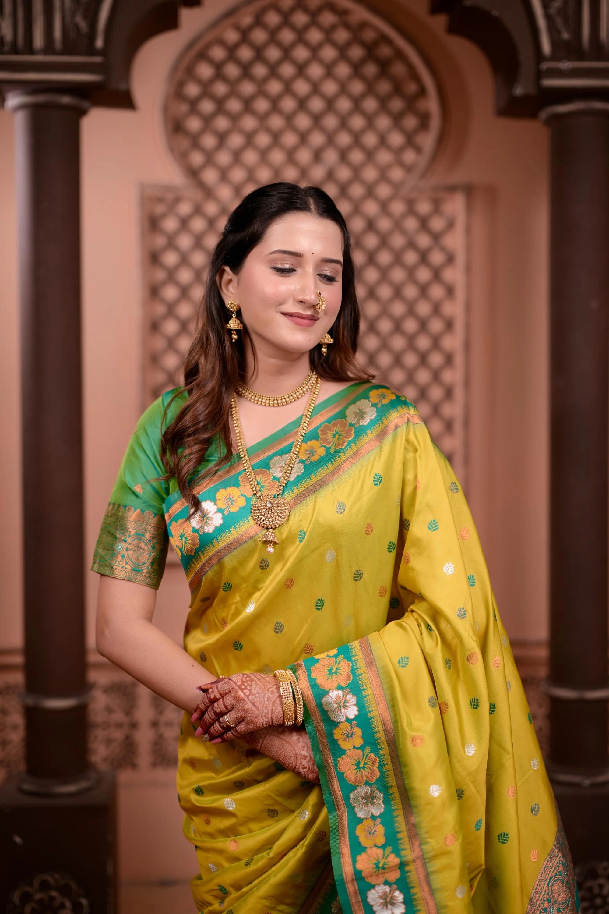 Woman in a yellow saree with green border standing in front of an ornate archway.