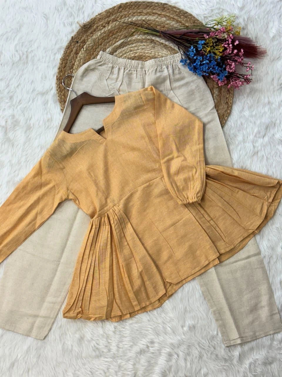 Mustard yellow dress with beige pants on a textured surface with a straw hat and flowers.