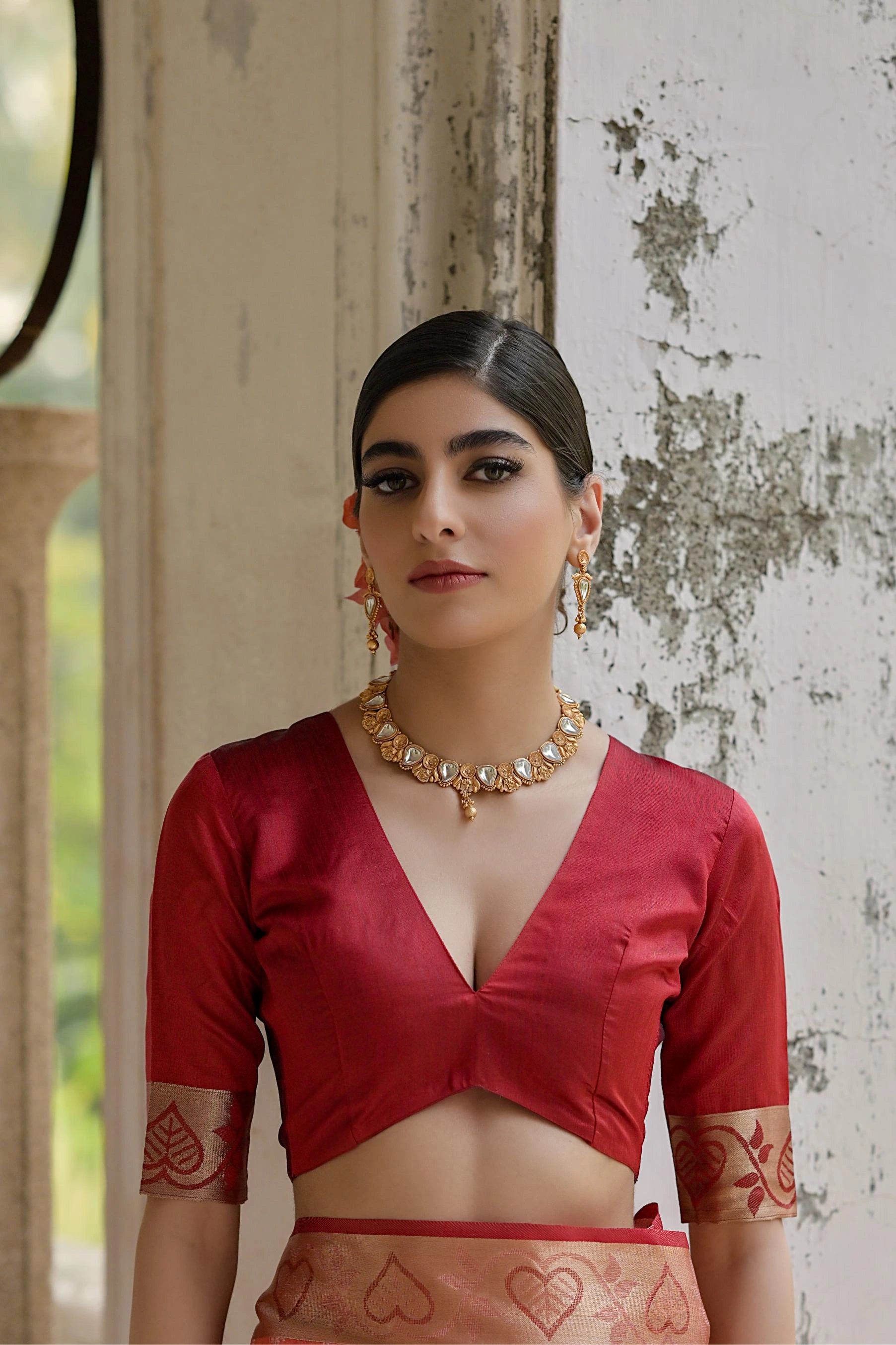 Woman wearing a red blouse with intricate designs, standing against a textured wall.