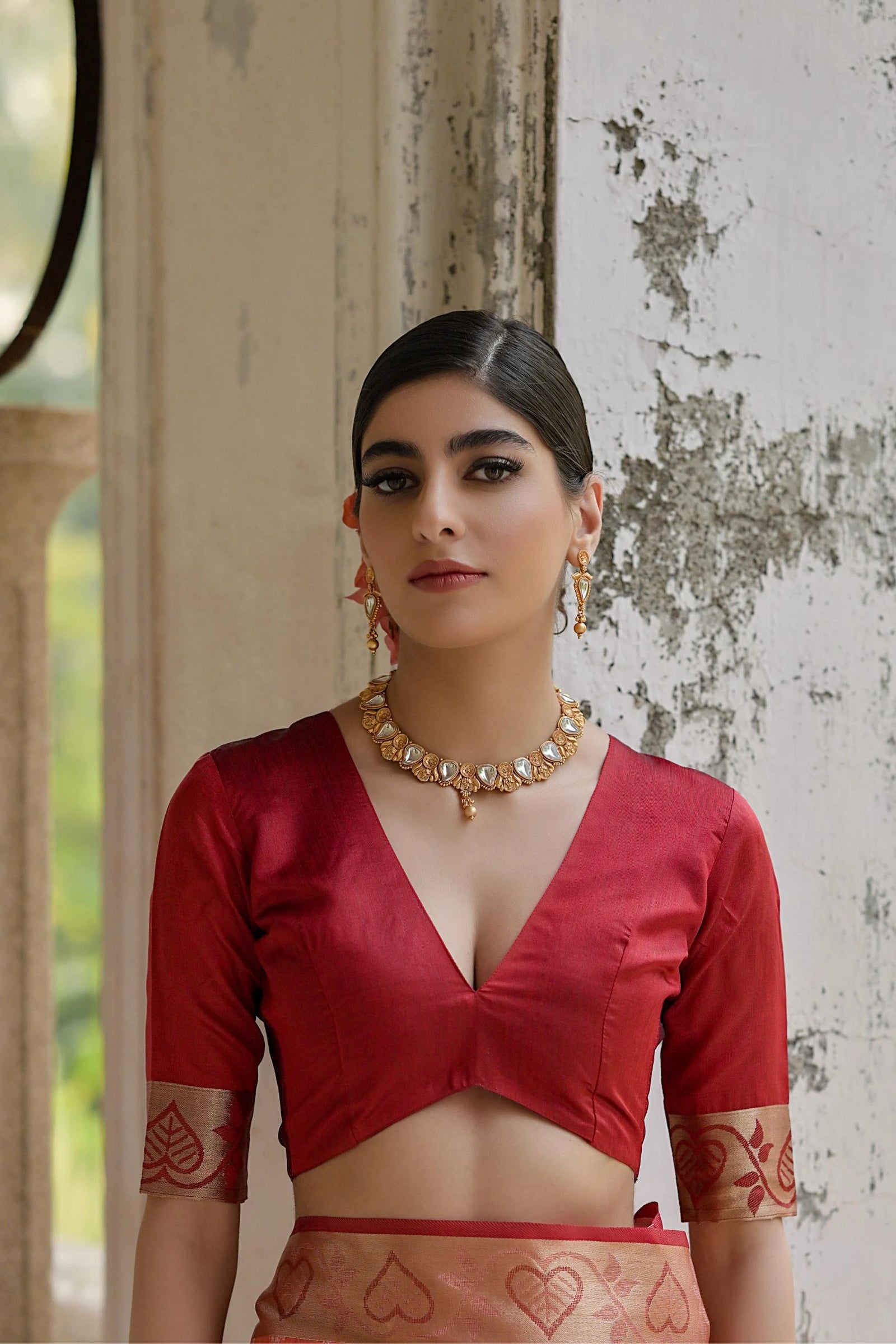 Woman wearing a red blouse with intricate designs, standing against a textured wall.