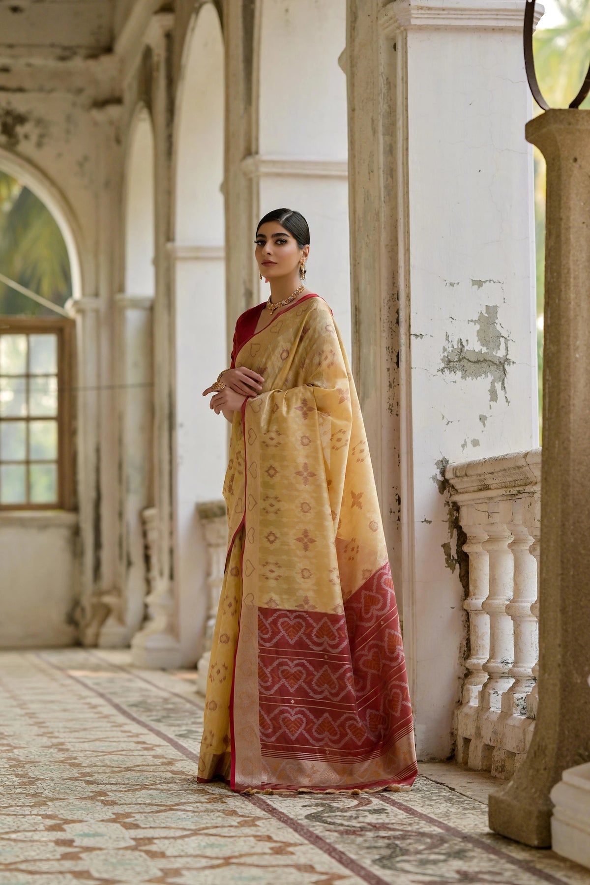 Woman in a yellow saree with a red border standing in an ornate room.