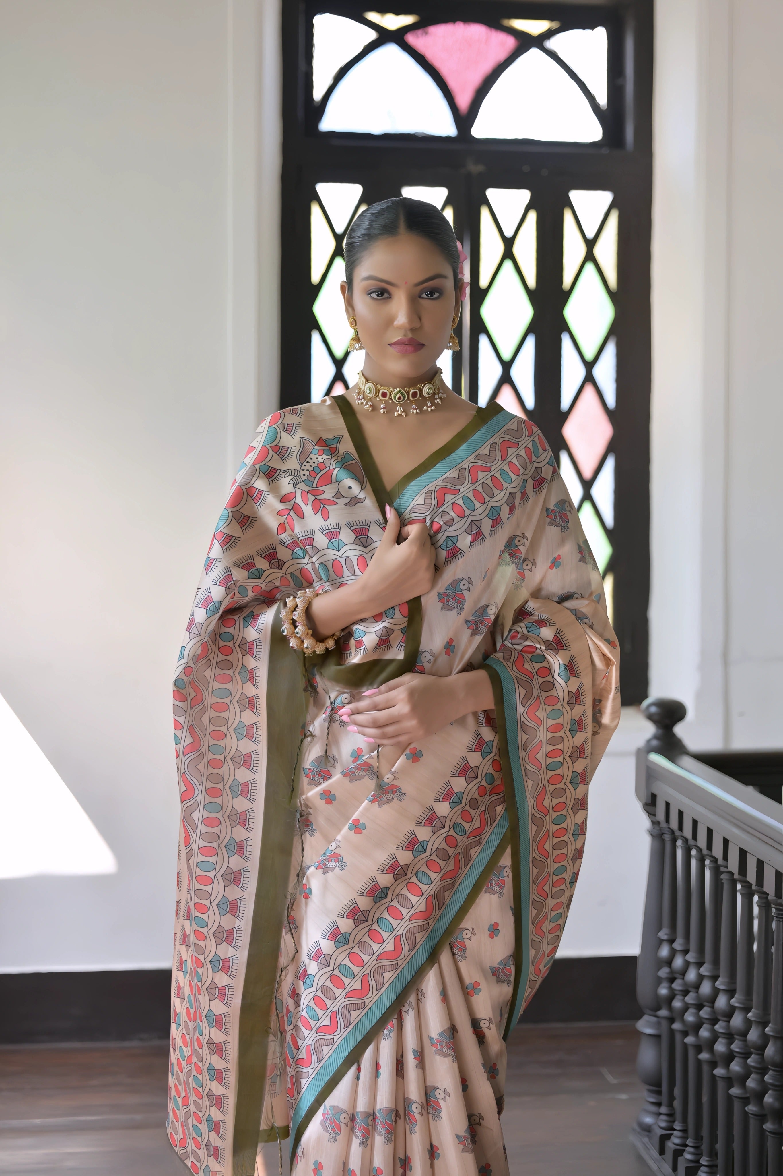 A woman models an elegant Beige Soft Tussar Silk Saree featuring an all-over Madhubani-style print in teal, coral, and green. The saree has a dark green and aqua border. She wears a contrasting olive green short-sleeve blouse and a gold Kundan choker necklace, posing indoors next to a dark wooden banister.