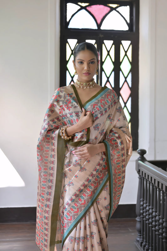 A woman models an elegant Beige Soft Tussar Silk Saree featuring an all-over Madhubani-style print in teal, coral, and green. The saree has a dark green and aqua border. She wears a contrasting olive green short-sleeve blouse and a gold Kundan choker necklace, posing indoors next to a dark wooden banister.