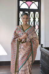 A woman models an elegant Beige Soft Tussar Silk Saree featuring an all-over Madhubani-style print in teal, coral, and green. The saree has a dark green and aqua border. She wears a contrasting olive green short-sleeve blouse and a gold Kundan choker necklace, posing indoors next to a dark wooden banister.