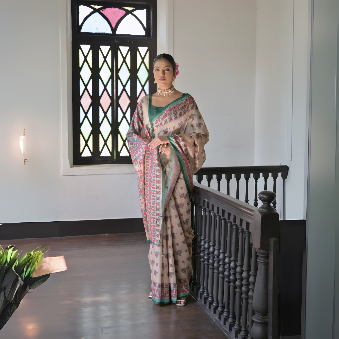 A woman models an elegant Off-White Soft Banarasi Silk Saree. The saree features a wide, prominent border and pallu with rich gold Zari weaving/print in shades of orange, peach, and green. She wears a coral sleeveless blouse and accessorizes with a gold Kundan choker necklace, large earrings, and a distinct dome-shaped handbag, posing indoors against a deep red backdrop.