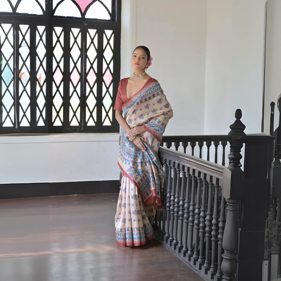 A woman models an elegant Beige Soft Tussar Silk Saree. The saree features an all-over Madhubani-style print of traditional motifs primarily in blue and rust/maroon. It is framed by a solid deep maroon border. She wears a deep maroon blouse and a gold choker necklace, posing indoors next to a dark wooden banister and window.