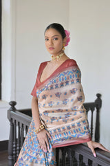 A woman models an elegant Beige Soft Tussar Silk Saree. The saree features an all-over Madhubani-style print of traditional motifs primarily in blue and rust/maroon. It is framed by a solid deep maroon border. She wears a deep maroon blouse and a gold choker necklace, posing indoors next to a dark wooden banister and window.
