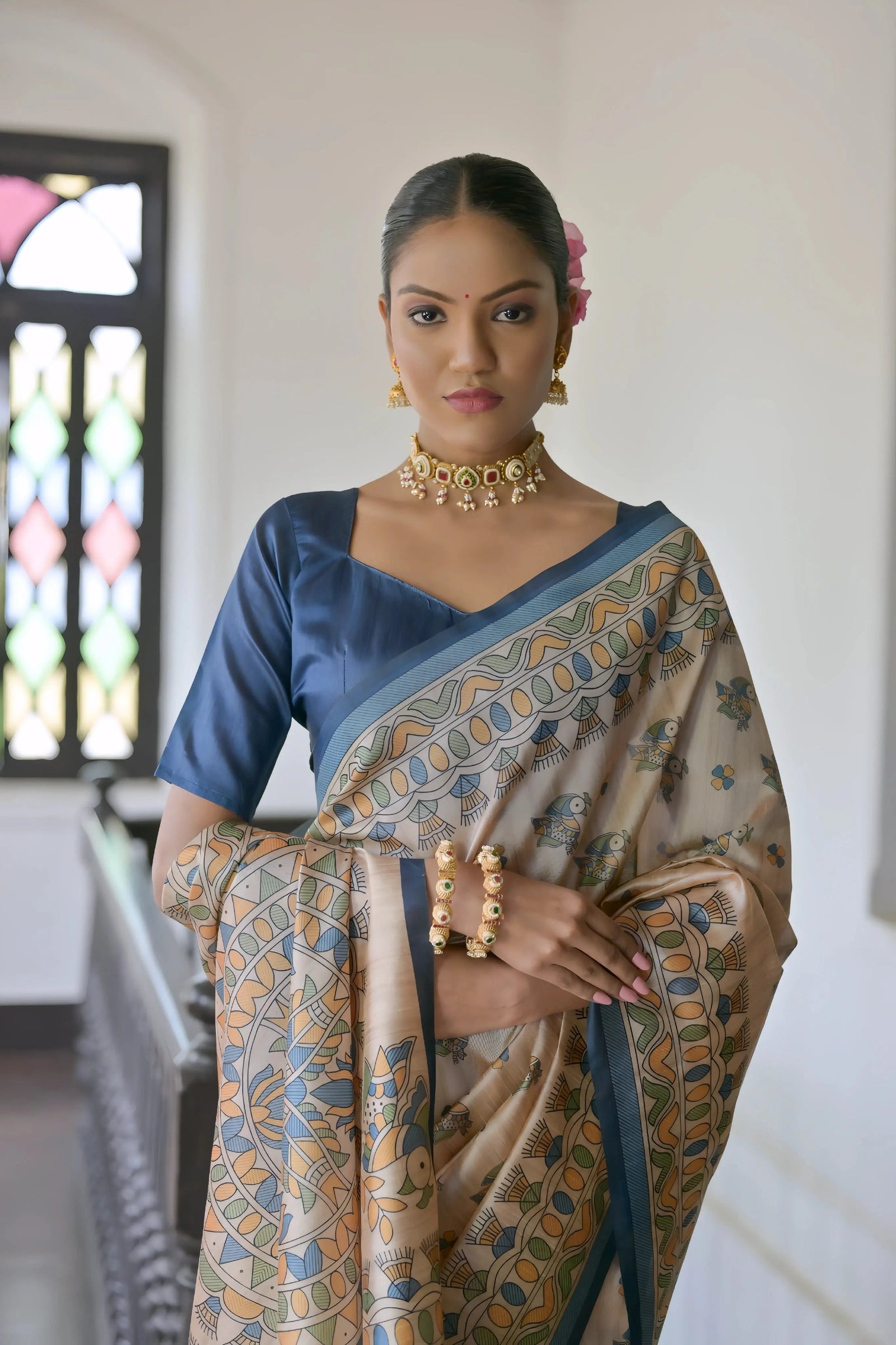 A woman models an elegant Beige Soft Tussar Silk Saree featuring an all-over Madhubani-style print in shades of blue and ochre. The saree is framed by a solid deep navy blue border. She wears a matching navy blue blouse and a gold Kundan choker necklace, posing indoors next to a dark wooden banister.