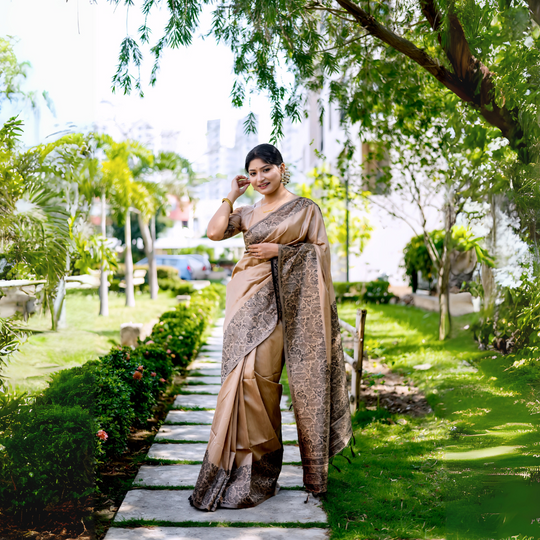 A woman models a luxurious Beige/Taupe Raw Silk Saree. The saree features a wide border and pallu with dense, intricate dark brown/black Zari weaving in a Kalamkari-style floral and paisley pattern. She wears a matching short-sleeve beige blouse and traditional gold jewelry, posing outdoors on a stone pathway.