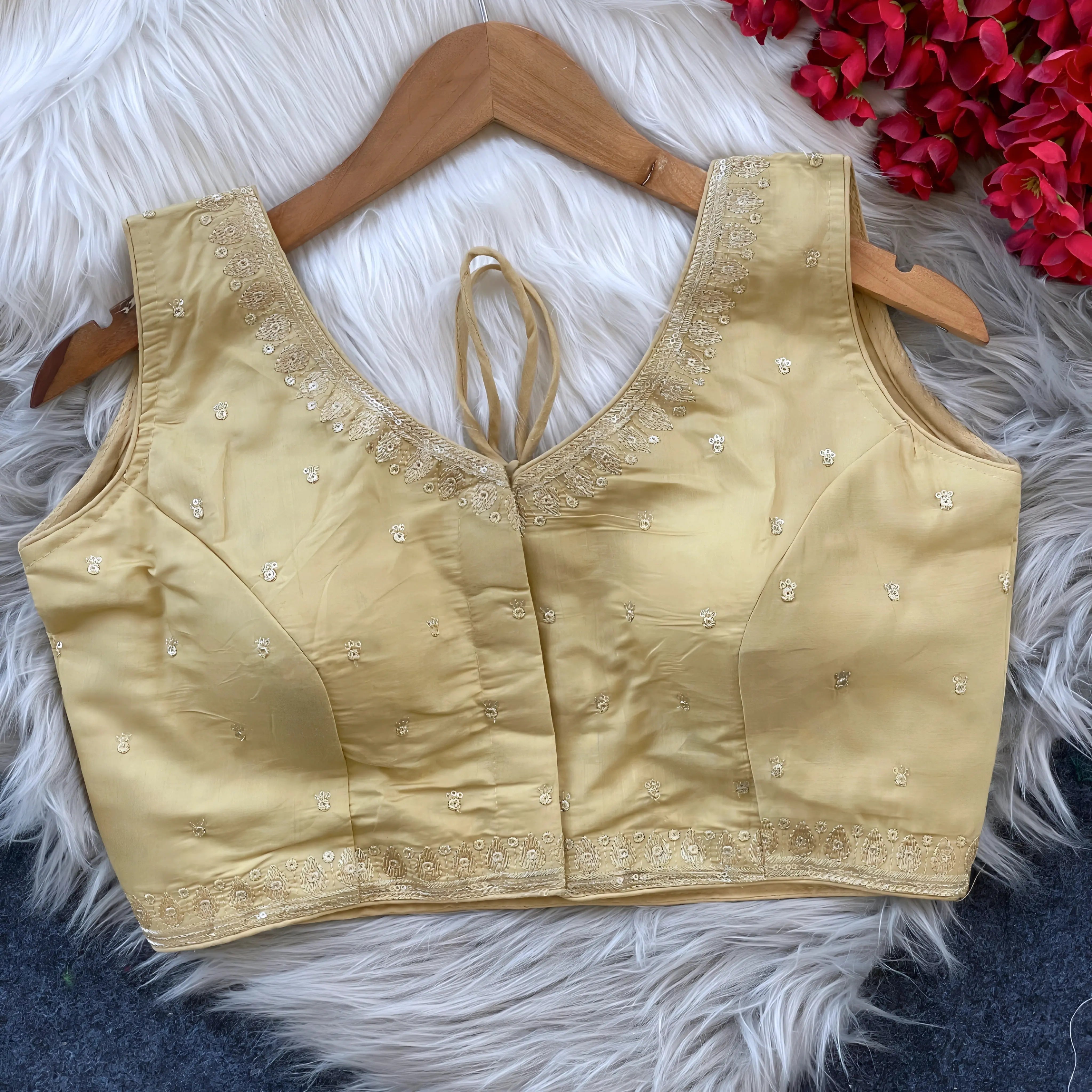  Beige padded blouse with delicate gold embroidery along the neckline and hem, featuring small sequin floral motifs, displayed on a wooden hanger over a white fur surface with red artificial flowers in the background.