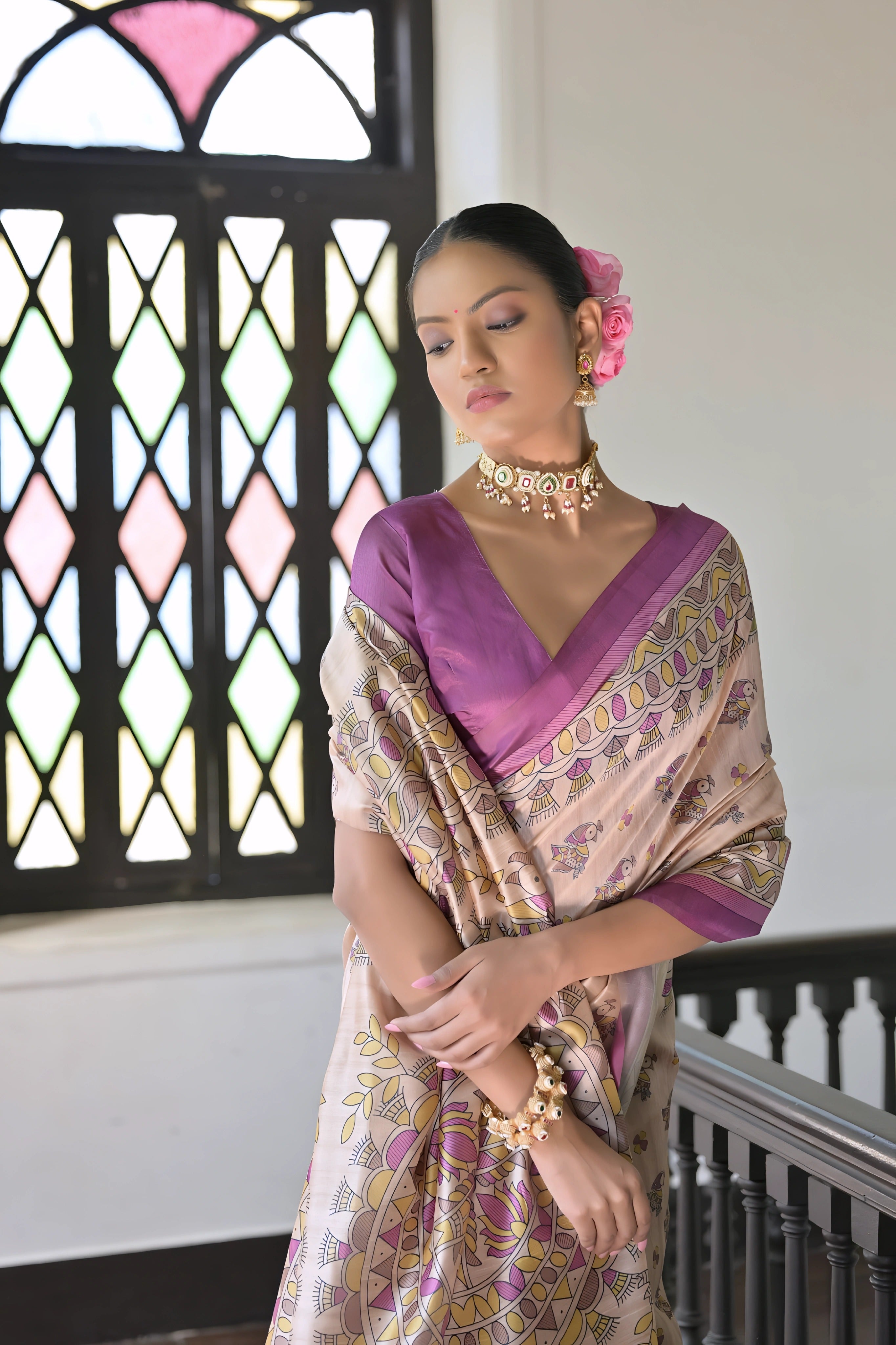 A woman models an elegant Beige Soft Tussar Silk Saree. The saree features an all-over Madhubani-style print of traditional motifs primarily in soft purple/lilac and yellow/green. It is framed by a vibrant deep purple/magenta border. She wears a matching purple sleeveless blouse and a light-toned choker necklace, posing indoors next to a dark wooden banister.