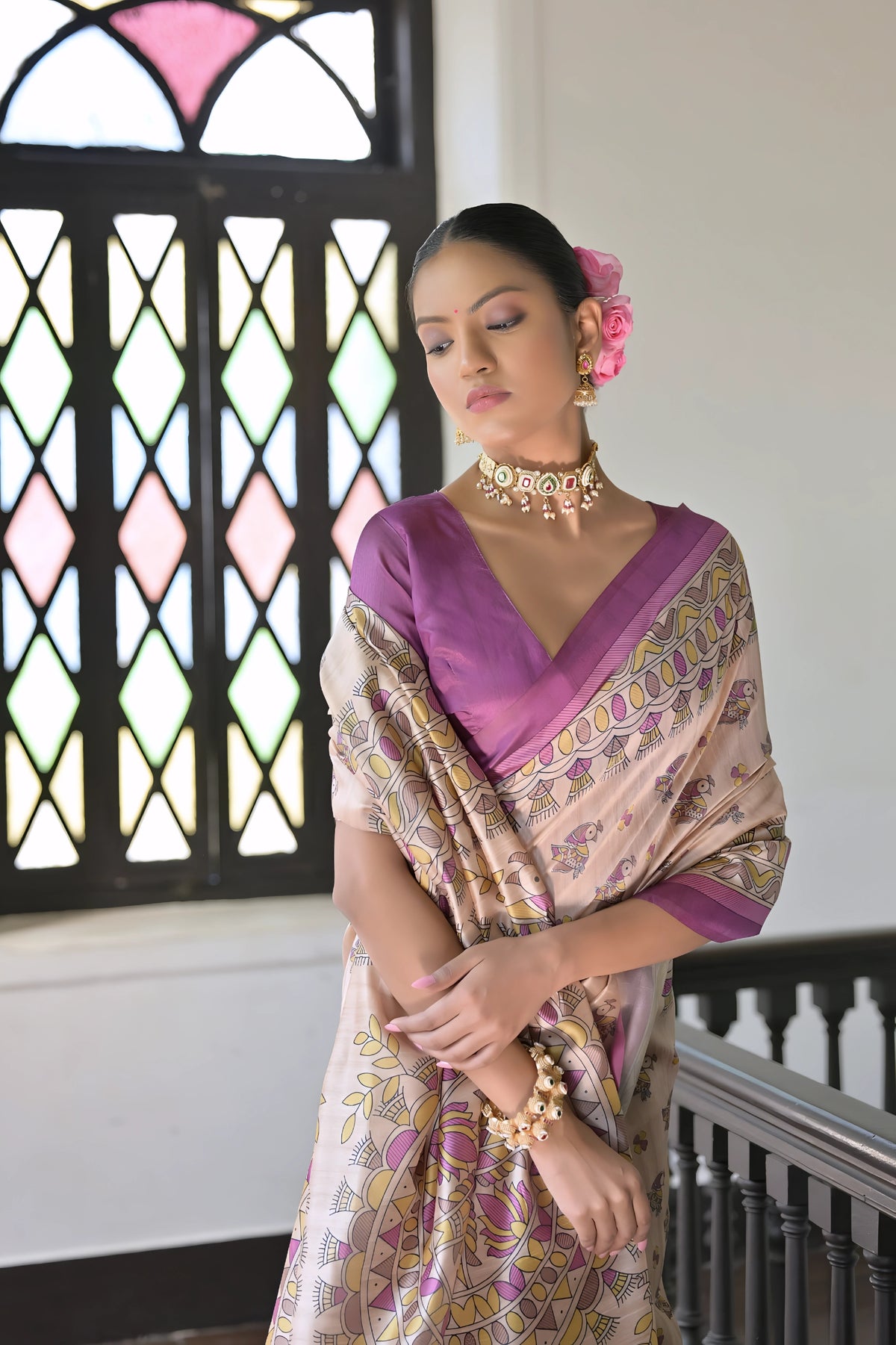 A woman models an elegant Beige Soft Tussar Silk Saree. The saree features an all-over Madhubani-style print of traditional motifs primarily in soft purple/lilac and yellow/green. It is framed by a vibrant deep purple/magenta border. She wears a matching purple sleeveless blouse and a light-toned choker necklace, posing indoors next to a dark wooden banister.
