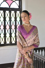A woman models an elegant Beige Soft Tussar Silk Saree. The saree features an all-over Madhubani-style print of traditional motifs primarily in soft purple/lilac and yellow/green. It is framed by a vibrant deep purple/magenta border. She wears a matching purple sleeveless blouse and a light-toned choker necklace, posing indoors next to a dark wooden banister.