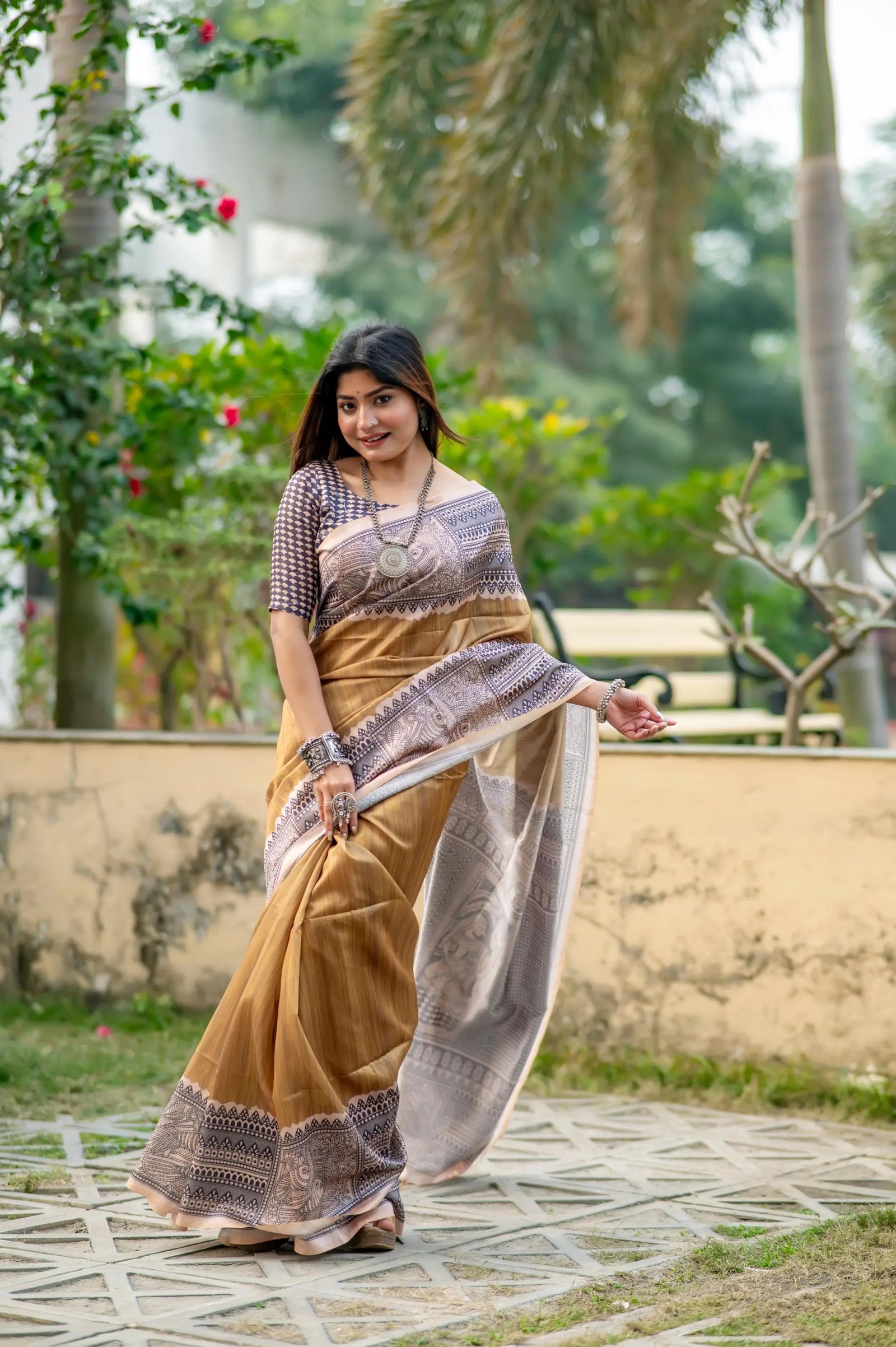 Woman in a beige saree with Madhubani print standing near a pond with lotus flowers in a lush garden setting
