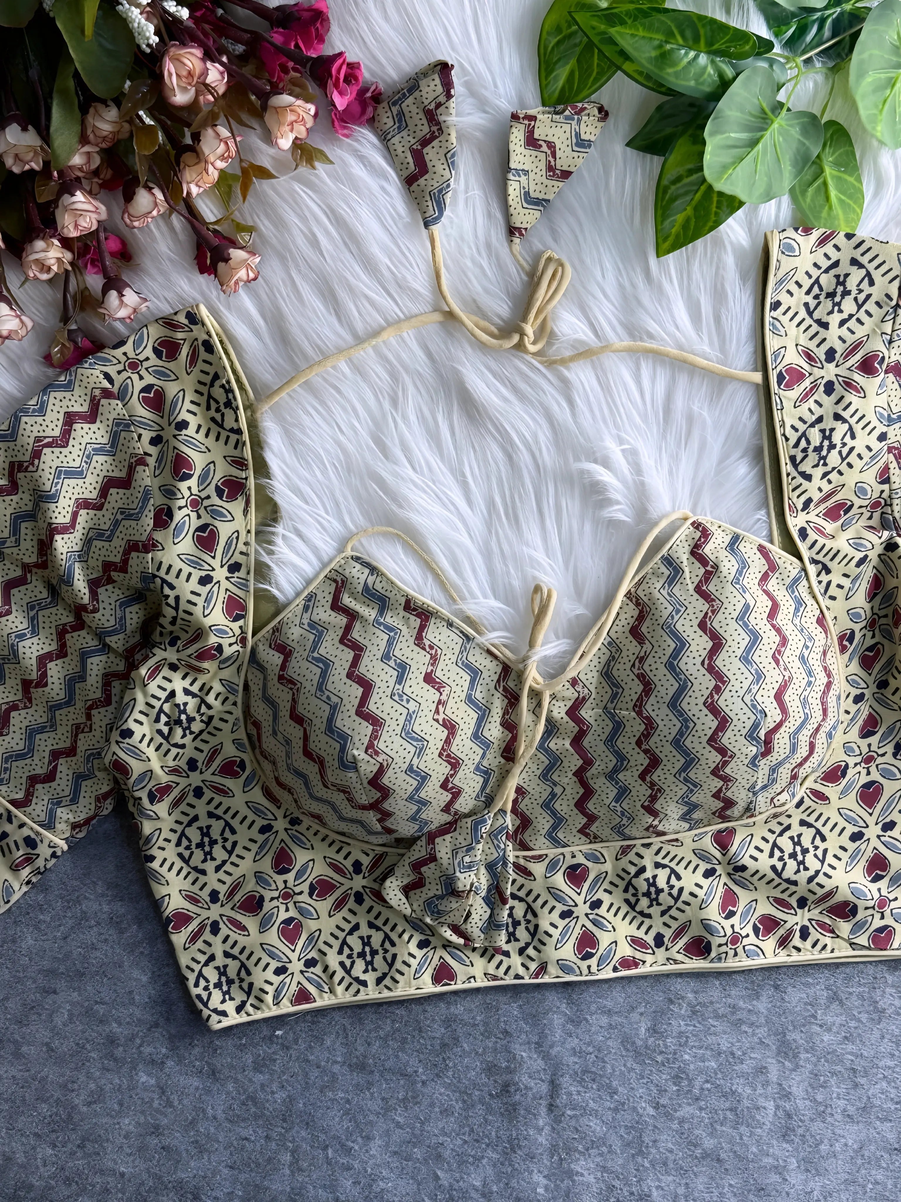 Beige padded blouse with maroon and navy geometric and floral patterns, displayed on a white fur surface with artificial flowers and green leaves in the background.