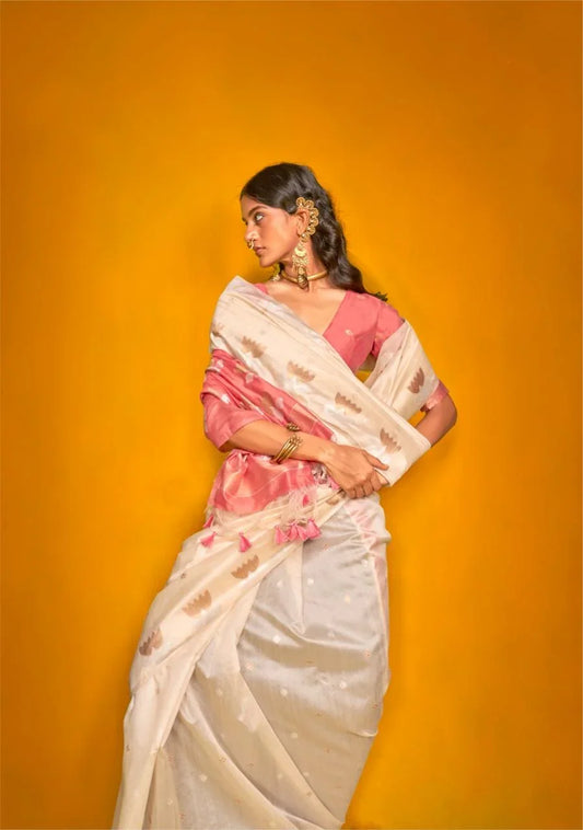 Woman wearing beige zari work silk saree with pink blouse and traditional jewellery posing against mustard yellow backdrop
