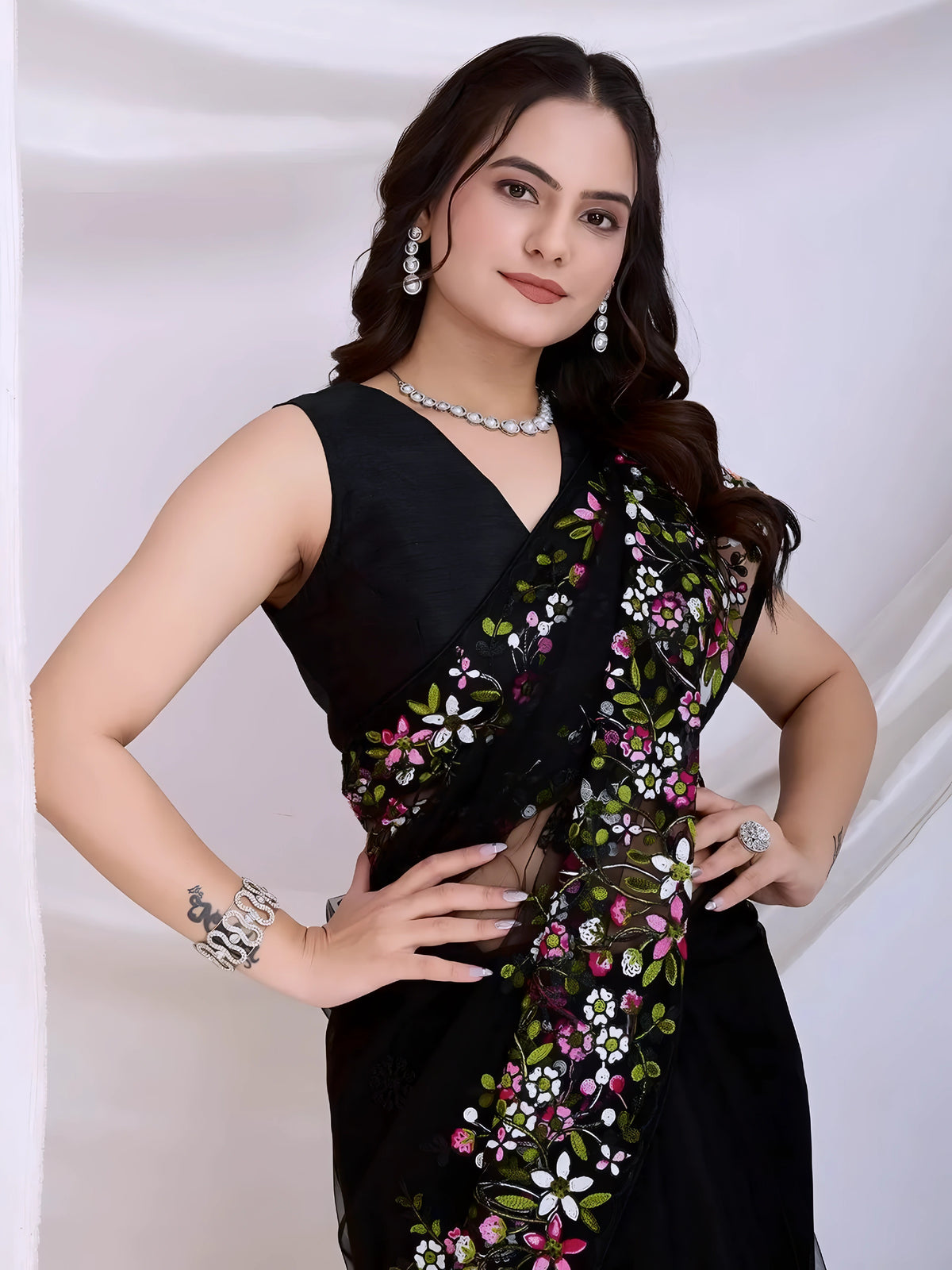 A woman models a dramatic Black Soft Net Saree. The sheer fabric is densely covered in vibrant all-over floral embroidery featuring rich pink, white, and green threadwork. She wears a matching black sleeveless blouse and accessorizes with sophisticated silver/diamond jewelry, posing indoors against sheer white curtains.