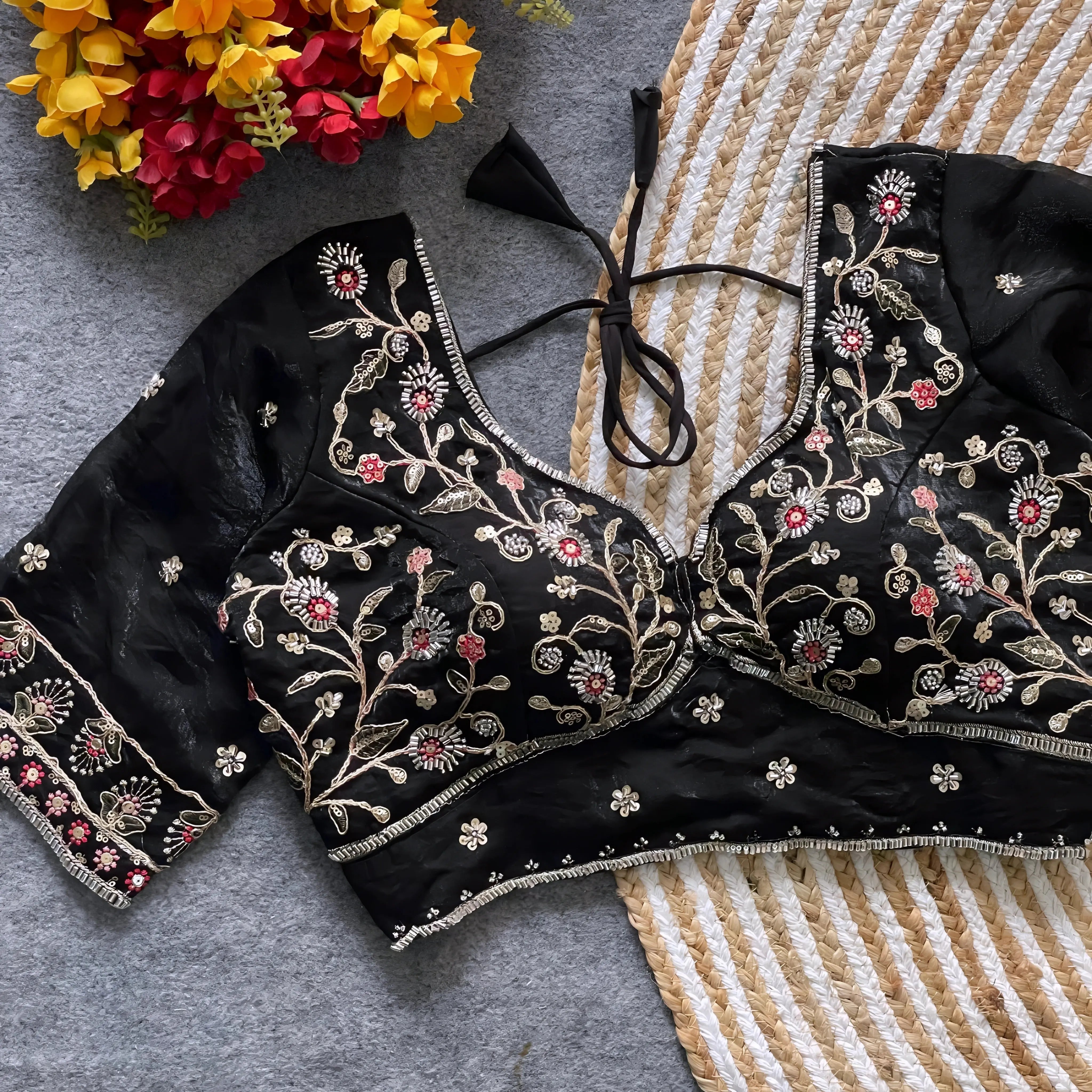 Black padded blouse with intricate floral embroidery in gold and red threads, featuring a tie-back detail, displayed on a grey surface with artificial yellow and red flowers and a woven mat in the background.