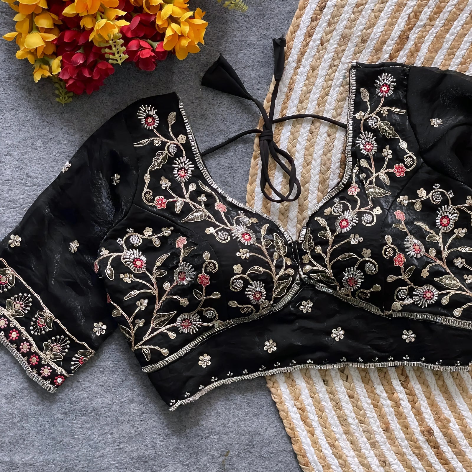 Black padded blouse with intricate floral embroidery in gold and red threads, featuring a tie-back detail, displayed on a grey surface with artificial yellow and red flowers and a woven mat in the background.