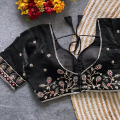 Black padded blouse with intricate floral embroidery in gold and red threads, featuring a tie-back detail, displayed on a grey surface with artificial yellow and red flowers and a woven mat in the background.