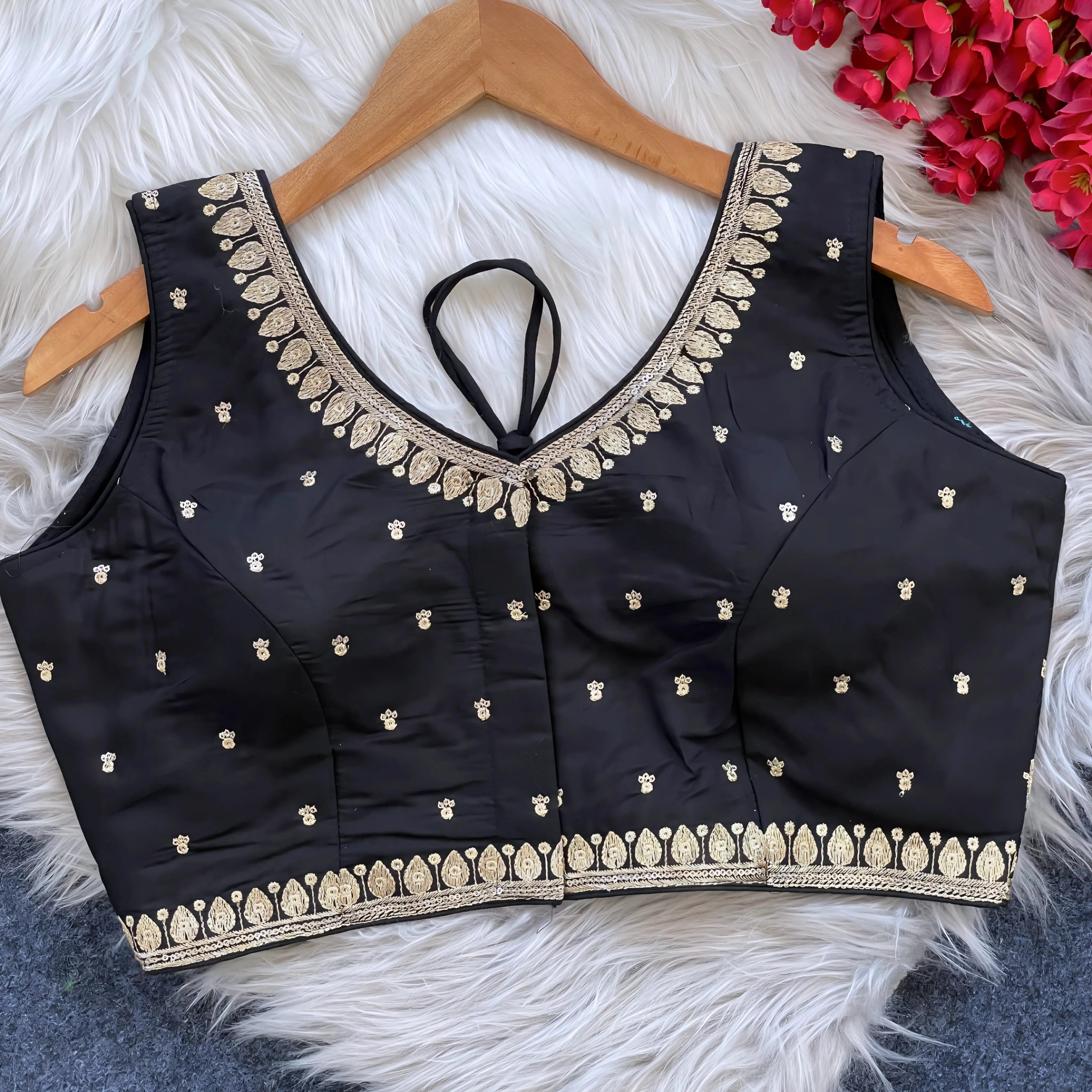Black padded blouse with intricate gold embroidery along the neckline and hem, featuring small gold embroidered motifs, displayed on a wooden hanger over a white fur surface with red artificial flowers in the background.
