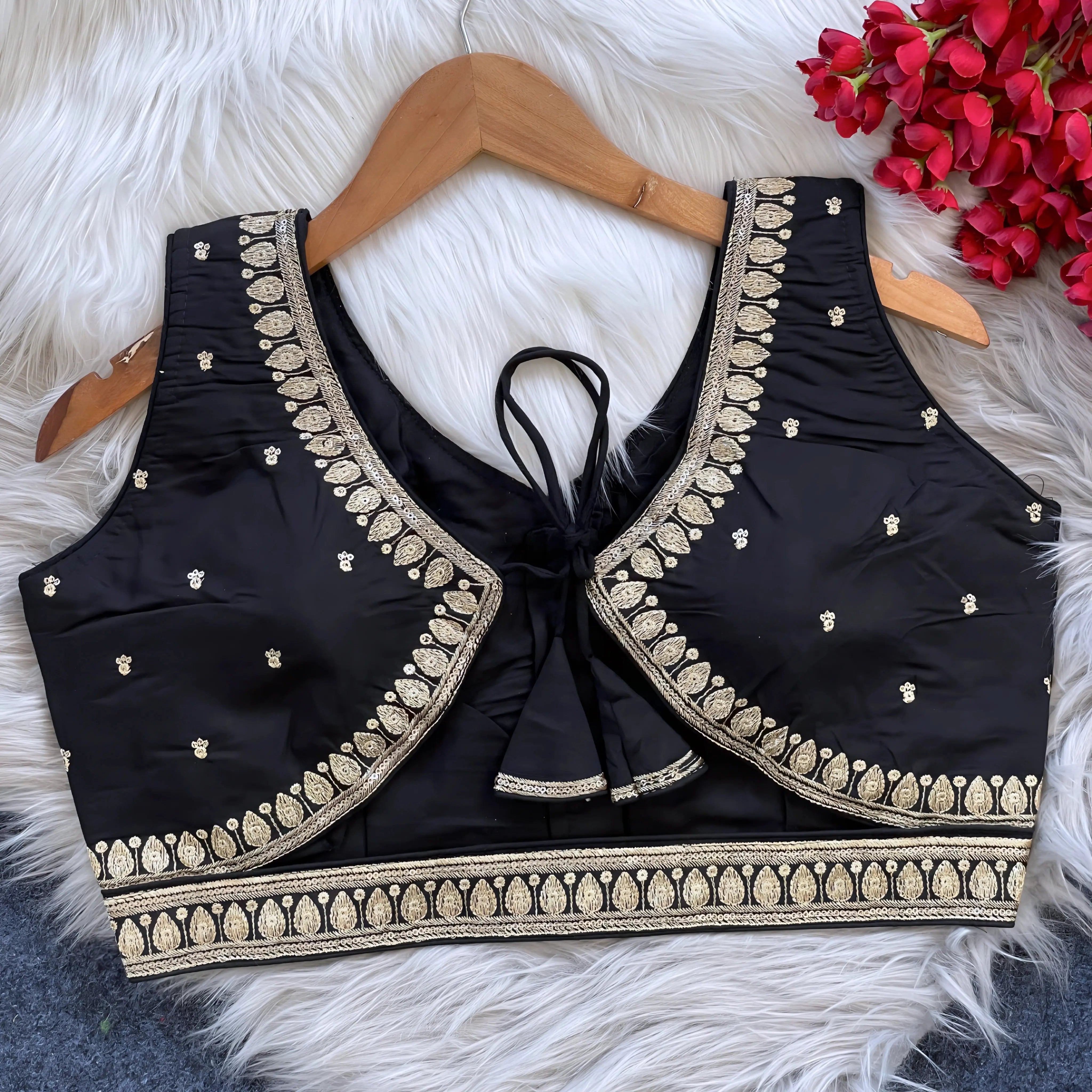 Black padded blouse with intricate gold embroidery along the neckline and hem, featuring small gold embroidered motifs, displayed on a wooden hanger over a white fur surface with red artificial flowers in the background.